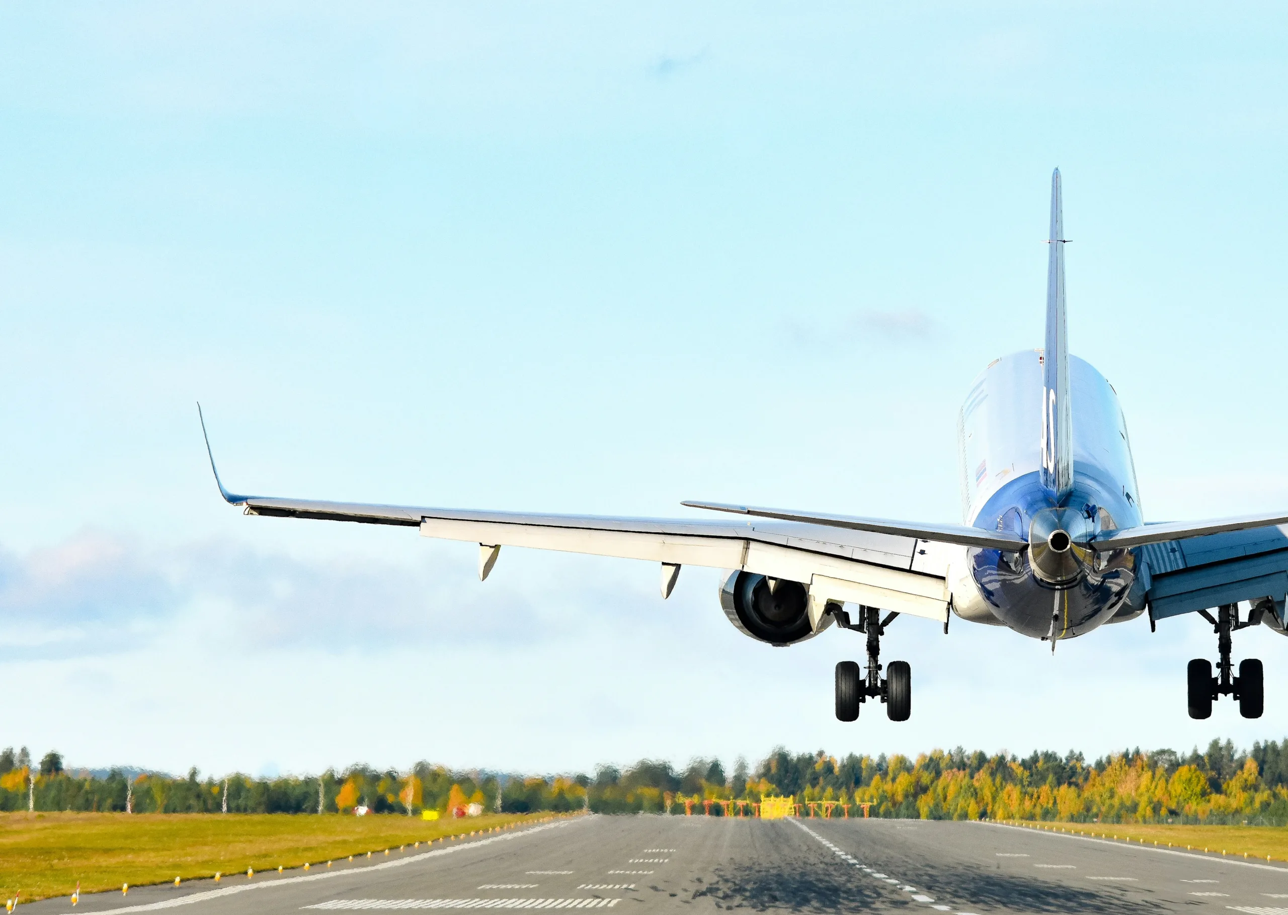 A commercial airplane taking off from a runway, with its landing gear still extended and trees lining the horizon under a bright sky.